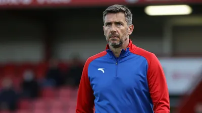 A concerned, mid-30s FC Halifax Town coach with a rugged build, wearing a plain blue and red training top, stands...