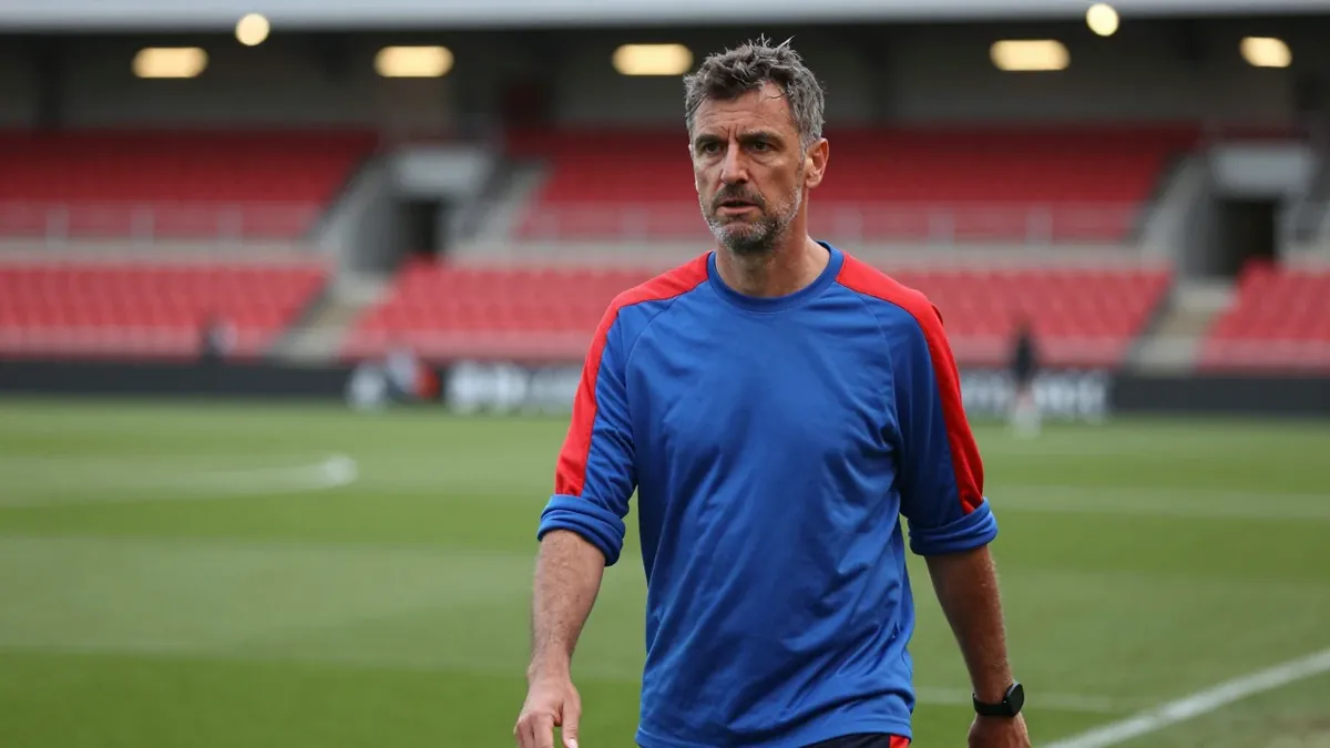 A tight, waist-up shot of a 35-year-old, ruggedly-built assistant coach, dressed in a plain, blue and red, cotton...