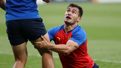 Mid-30s muscular forward, head tilted back, eyes wide, caught mid-tackle as a teammate's leg crashes into his torso...