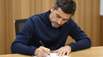 A 40-year-old male football manager in a navy blue tracksuit, head-and-shoulders, signing a contract at a wooden desk...
