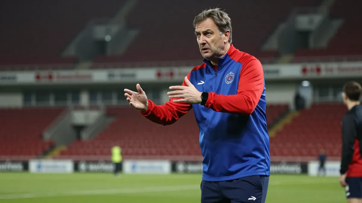 A 50-year-old male coach, dressed in a plain blue and red polyester training top, stands on the pitchside grass at The...