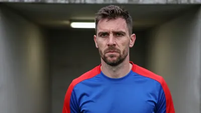A close-up shot of a 35-year-old male coach with a weathered face, standing in the tunnel of The Shay Stadium, wearing a...