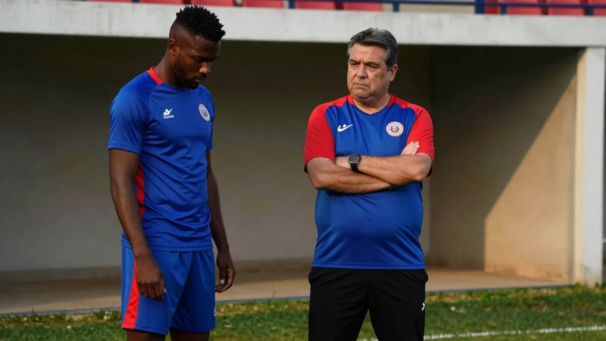 A 50-year-old, stocky, dark-haired coach, dressed in a plain blue and red polyester training top, stands sternly on the...