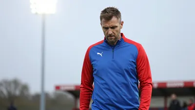 A concerned, rugged, 30-something FC Halifax Town coach, in a plain blue and red training top, stands pitchside, soft...