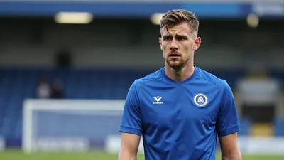 A 30-year-old FC Halifax Town player, wearing a solid blue kit, stands in front of the Shay Stadium, looking concerned
