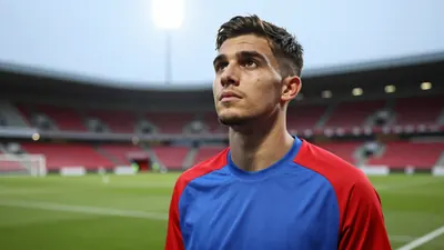 A close-up shot of a 28-year-old, 5'10" midfielder, dressed in a plain blue and red polyester training top, standing at...
