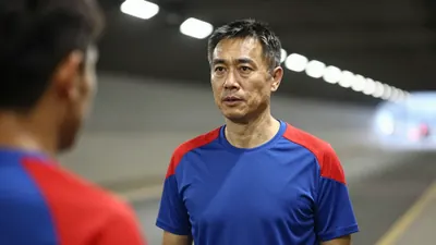 A 50-year-old, 5'10" male coach, wearing a plain blue and red polyester training top, stands intensely in the low-haze of...