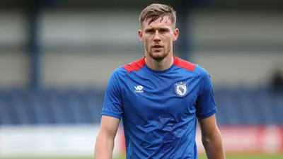 A determined, rugged, 28-year-old FC Halifax Town midfielder, wearing a plain blue and red training top, stands on the...
