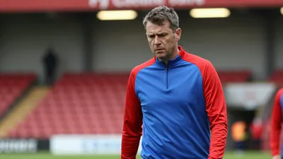A concerned, mid-30s FC Halifax Town coach, with a rugged build, wearing a plain, unmarked blue and red training top...