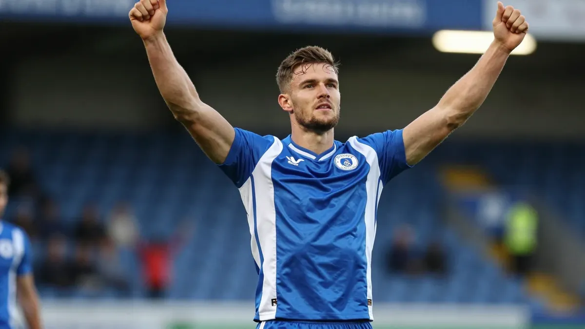 A 30-year-old FC Halifax Town player, with a muscular build, in a blue and white kit, celebrating a goal with his arms...