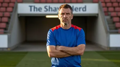 A 35-year-old male coach in a plain blue and red polyester training top, his weathered face set in a determined...