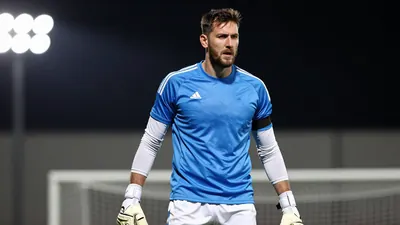 A 35-year-old, tall, athletic goalkeeper in a blue jersey with white sleeves and white shorts, wearing a white pair of...