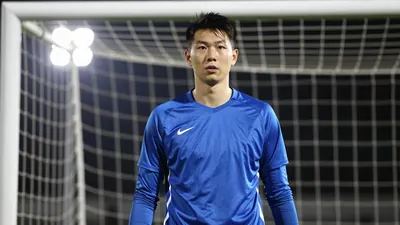 A 25-year-old goalkeeper in a solid blue kit, standing in a goalmouth with a determined expression