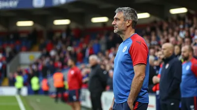 A 35-year-old male assistant coach, with a rugged build and graying temples, stands at the edge of the pitch, gazing out...