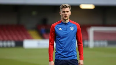 A determined 25-year-old FC Halifax Town midfielder, wearing a plain blue and red training top, stands on the pitchside...