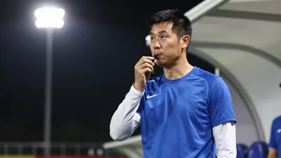 A 28-year-old male assistant coach in a blue jersey with white sleeves, standing on the touchline with a whistle in hand