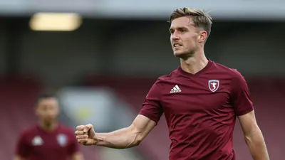 A 25-year-old FC Halifax Town midfielder, athletic build, in solid maroon kit, celebrates scoring with a clenched fist